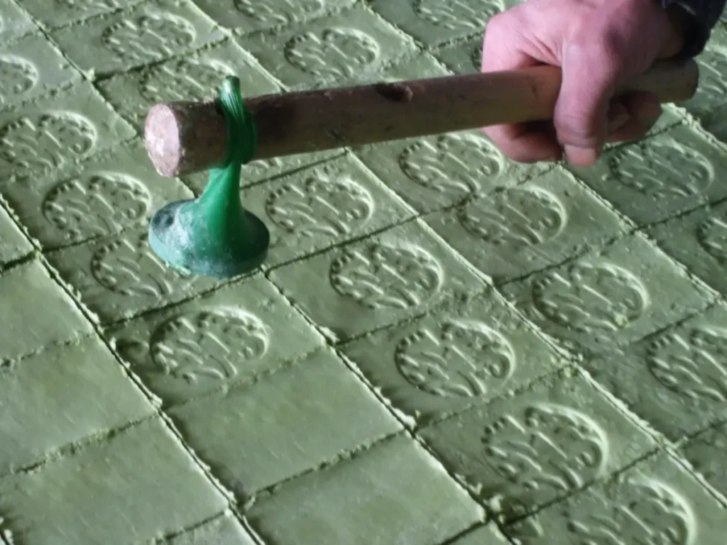 A close-up of an artisan's hand using a traditional wooden hammer and stamp to imprint a producer's seal onto fresh green Aleppo soap blocks.