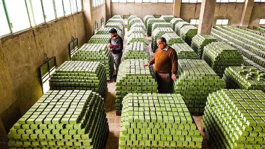 Two men inspecting thousands of hand-cut blocks of fresh green Aleppo laurel soap stacked in a large, sunlit factory warehouse in Syria.