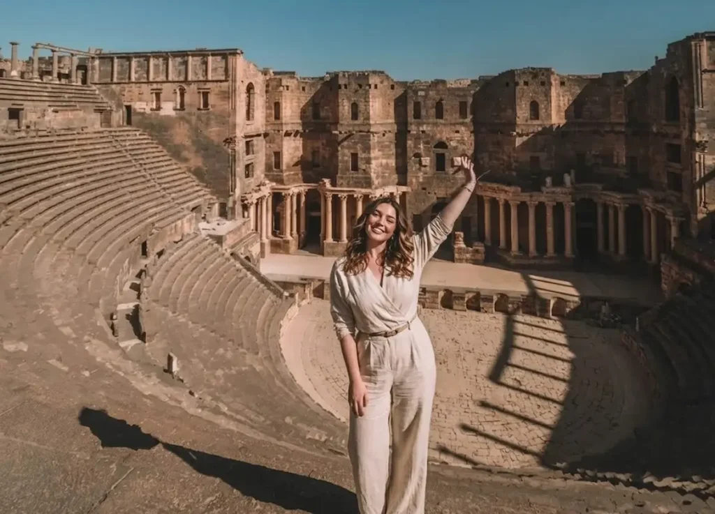 A wide-angle view of a woman in a beige jumpsuit standing with one arm raised in the ancient Roman Theater of Bosra, Syria, with the massive semi-circular stone seating and the ornate stage backdrop visible behind her.