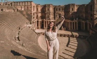 A wide-angle view of a woman in a beige jumpsuit standing with one arm raised in the ancient Roman Theater of Bosra, Syria, with the massive semi-circular stone seating and the ornate stage backdrop visible behind her.