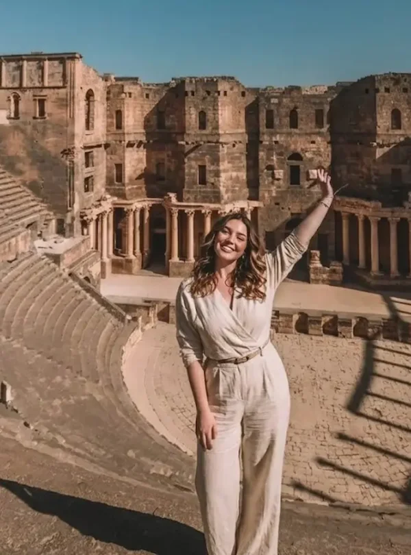 A wide-angle view of a woman in a beige jumpsuit standing with one arm raised in the ancient Roman Theater of Bosra, Syria, with the massive semi-circular stone seating and the ornate stage backdrop visible behind her.