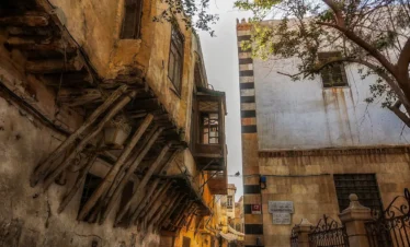 A vertical shot of a narrow cobblestone street in Old Damascus during golden hour, featuring historic multi-story buildings and traditional wooden supports.