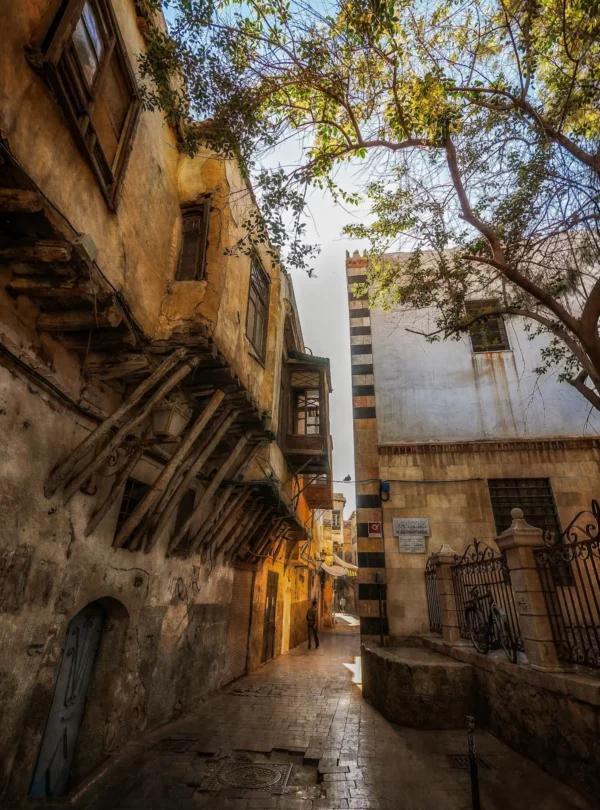 A vertical shot of a narrow cobblestone street in Old Damascus during golden hour, featuring historic multi-story buildings and traditional wooden supports.