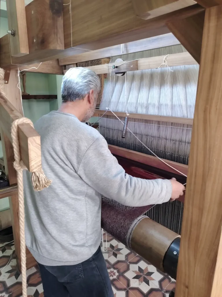 A man in a grey sweater seen from behind, operating a large, traditional wooden hand-loom to weave a dark purple fabric with intricate floral brocade patterns in a workshop.