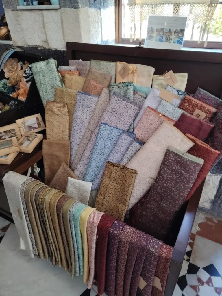 A large wooden chest filled with dozens of folded rolls of traditional Damascene brocade fabric in various colors like gold, blue, burgundy, and silver, each featuring intricate floral and bird patterns.