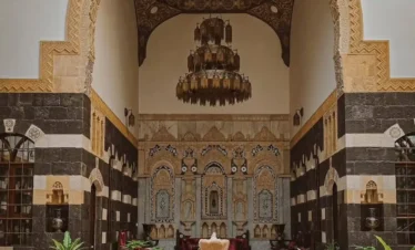 An ornate courtyard of a traditional Damascene house featuring a grand stone archway (Iwan) with a vaulted wooden ceiling, an intricate crystal chandelier, and walls decorated in the bicolor "Ablaq" style with stone carvings. A stone fountain and lush green plants sit in the foreground.