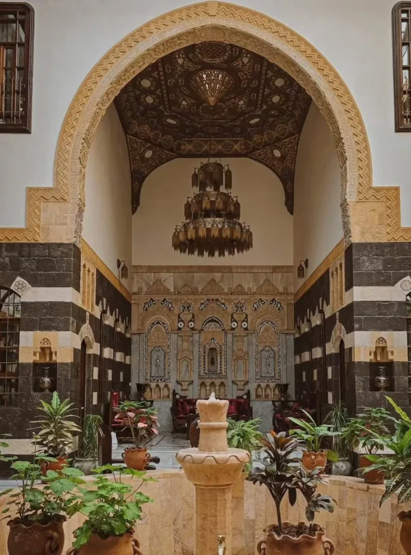 An ornate courtyard of a traditional Damascene house featuring a grand stone archway (Iwan) with a vaulted wooden ceiling, an intricate crystal chandelier, and walls decorated in the bicolor "Ablaq" style with stone carvings. A stone fountain and lush green plants sit in the foreground.