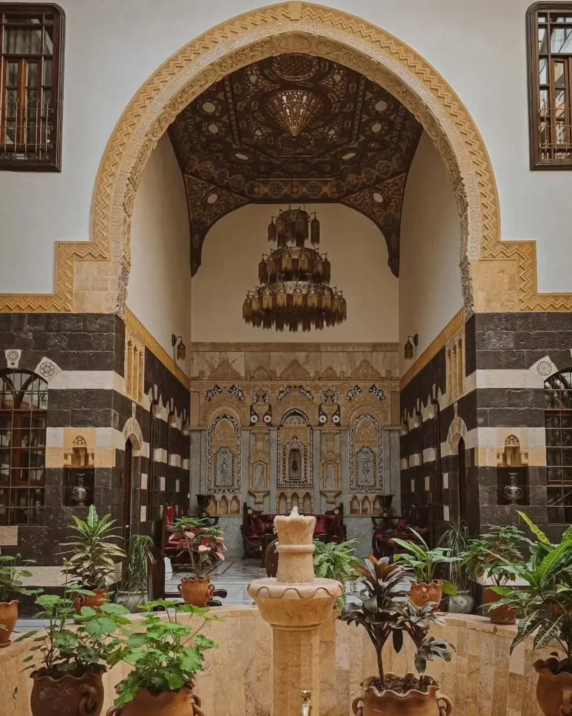 An ornate courtyard of a traditional Damascene house featuring a grand stone archway (Iwan) with a vaulted wooden ceiling, an intricate crystal chandelier, and walls decorated in the bicolor "Ablaq" style with stone carvings. A stone fountain and lush green plants sit in the foreground.
