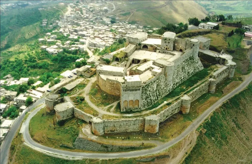 An aerial view of the massive stone Krak des Chevaliers crusader castle perched on a lush green hilltop in Homs, Syria, overlooking a small village and rolling valley.