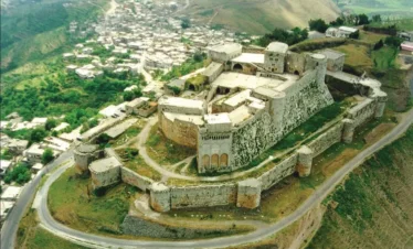 An aerial view of the massive stone Krak des Chevaliers crusader castle perched on a lush green hilltop in Homs, Syria, overlooking a small village and rolling valley.