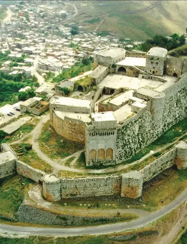 An aerial view of the massive stone Krak des Chevaliers crusader castle perched on a lush green hilltop in Homs, Syria, overlooking a small village and rolling valley.
