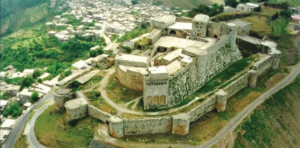An aerial view of the massive stone Krak des Chevaliers crusader castle perched on a lush green hilltop in Homs, Syria, overlooking a small village and rolling valley.