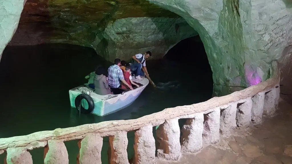 A small white boat carrying several passengers through a large underground limestone cavern with an emerald-green subterranean lake, guided by a man using a wooden pole.