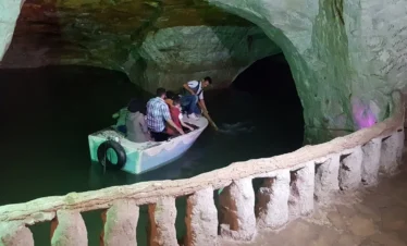 A small white boat carrying several passengers through a large underground limestone cavern with an emerald-green subterranean lake, guided by a man using a wooden pole.