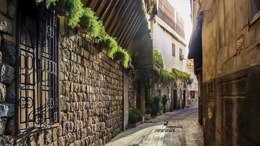 A narrow, sun-drenched cobblestone street in Old Damascus lined with ancient stone buildings featuring wrought-iron window grilles, overhanging wooden eaves, and vibrant green plants cascading from balconies.