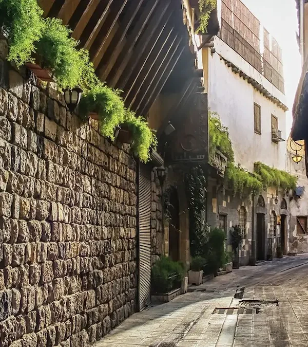 A narrow, sun-drenched cobblestone street in Old Damascus lined with ancient stone buildings featuring wrought-iron window grilles, overhanging wooden eaves, and vibrant green plants cascading from balconies.