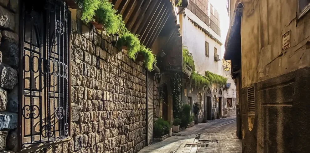 A narrow, sun-drenched cobblestone street in Old Damascus lined with ancient stone buildings featuring wrought-iron window grilles, overhanging wooden eaves, and vibrant green plants cascading from balconies.