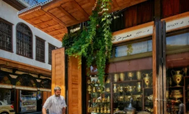 A street-level view in Old Damascus showing a man with a bicycle walking past a wooden-fronted shop filled with polished gold and brass vessels. A large, ornate traditional coffee pot (Dallah) sits on the cobblestone pavement in front.