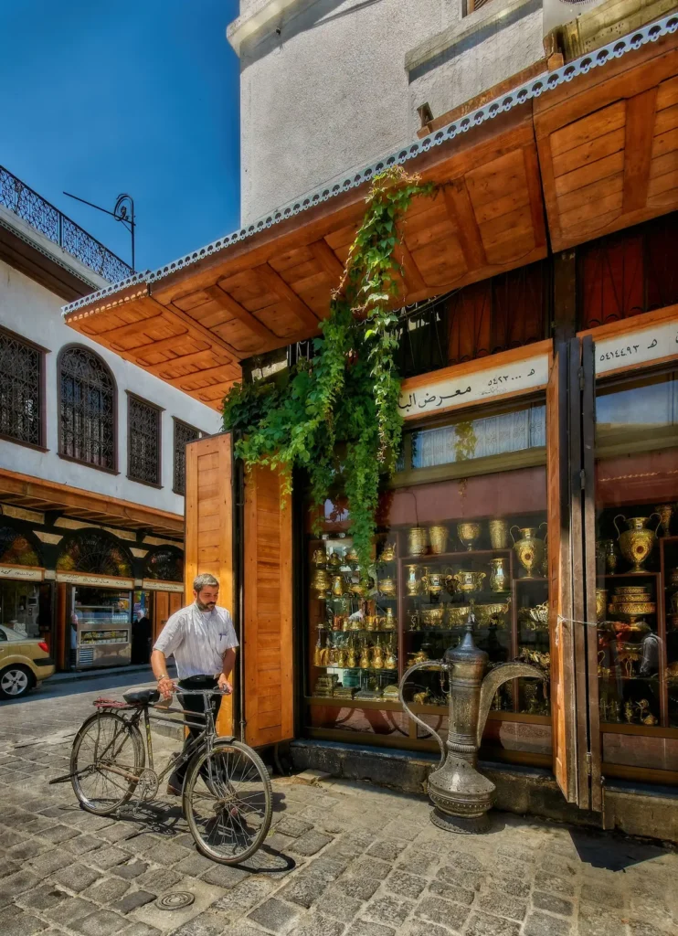 A street-level view in Old Damascus showing a man with a bicycle walking past a wooden-fronted shop filled with polished gold and brass vessels. A large, ornate traditional coffee pot (Dallah) sits on the cobblestone pavement in front.