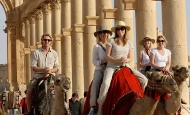 Three camels carrying tourists through the sun-drenched ruins of Palmyra, Syria, passing beside a long row of towering ancient Roman columns.