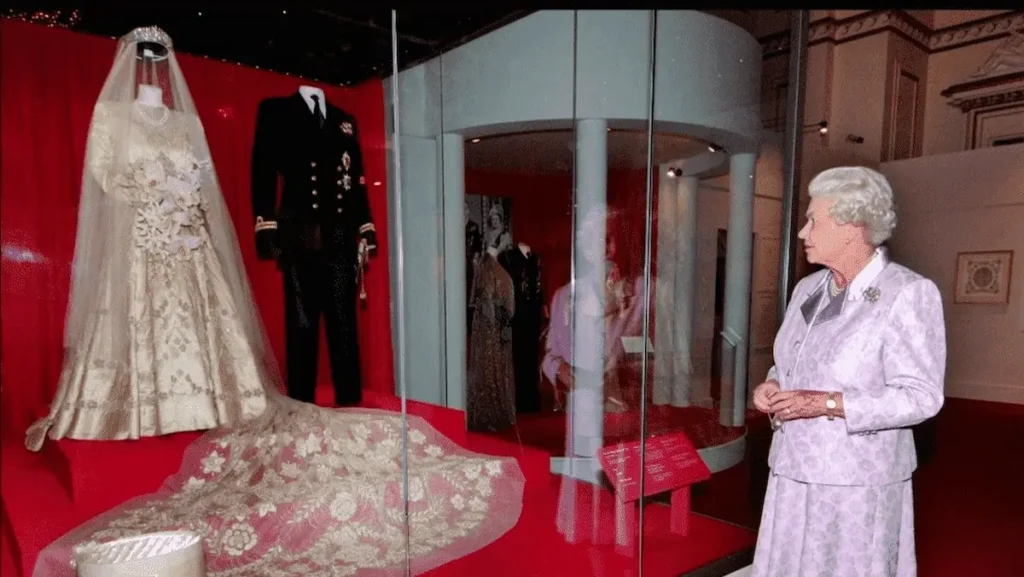 Queen Elizabeth II standing in a museum gallery, looking at a display case containing her 1947 wedding gown and a naval uniform. The gown is made of ornate ivory Damascene brocade with intricate floral patterns.