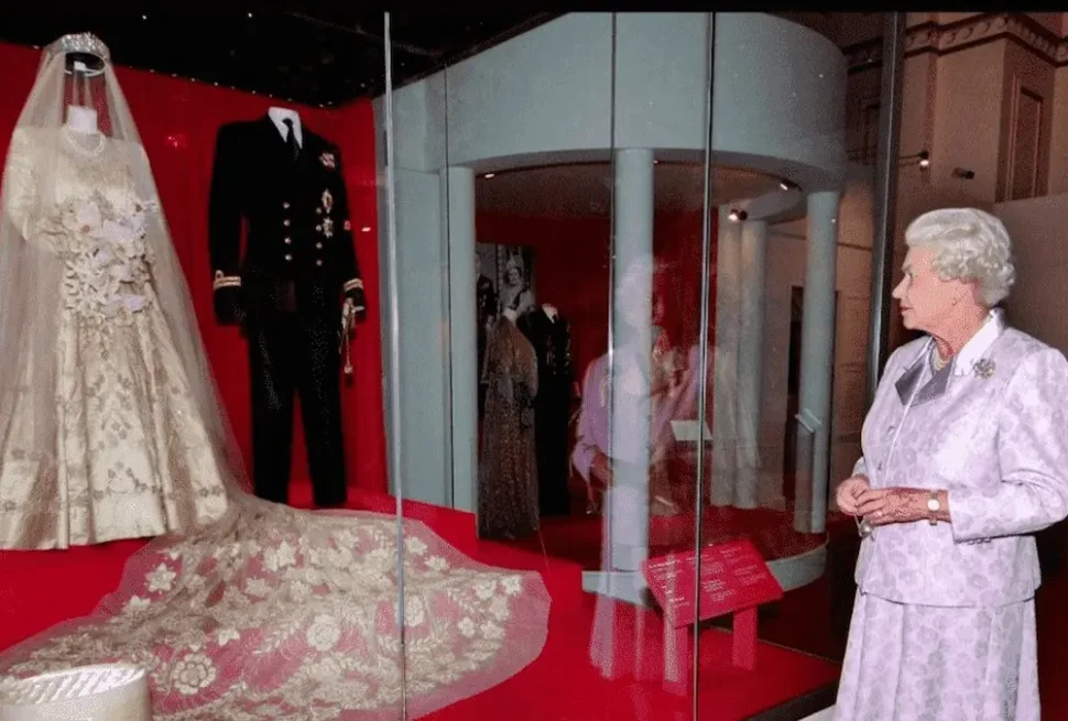 Queen Elizabeth II standing in a museum gallery, looking at a display case containing her 1947 wedding gown and a naval uniform. The gown is made of ornate ivory Damascene brocade with intricate floral patterns.