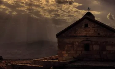 A dramatic silhouette of a stone chapel at the Cherubim Monastery in Saidnaya, Syria, under a bright sun breaking through dark, textured clouds over a mountainous horizon.