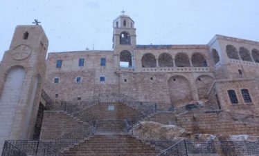 The historic stone facade and bell tower of the Convent of Our Lady in Saidnaya, Syria, covered in light snow with intricate stairs leading to the entrance.