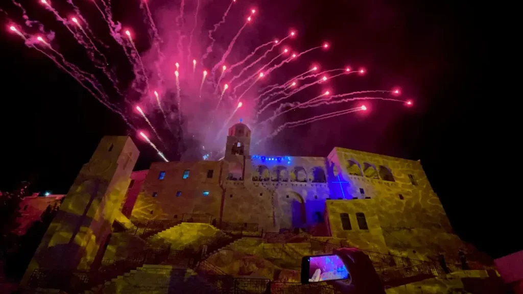 A night photograph taken from a low angle showing a smartphone recording pink and white fireworks exploding over the illuminated stone facade of the Our Lady of Saidnaya Monastery.