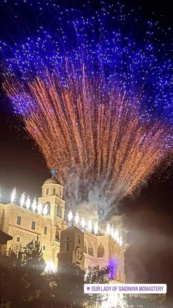 A night photograph of Our Lady of Saidnaya Monastery in Syria illuminated by a large display of blue and orange fireworks during a festival.