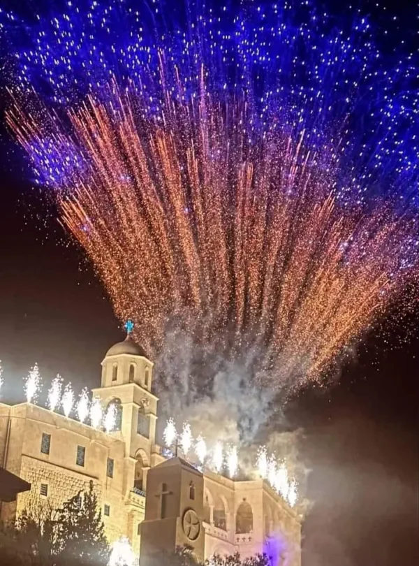 A night photograph of Our Lady of Saidnaya Monastery in Syria illuminated by a large display of blue and orange fireworks during a festival.
