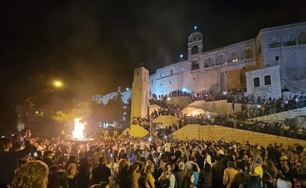 A large crowd of people gathered at night in front of the illuminated stone walls of the Our Lady of Saidnaya Monastery, watching a large ceremonial bonfire during a traditional religious festival.