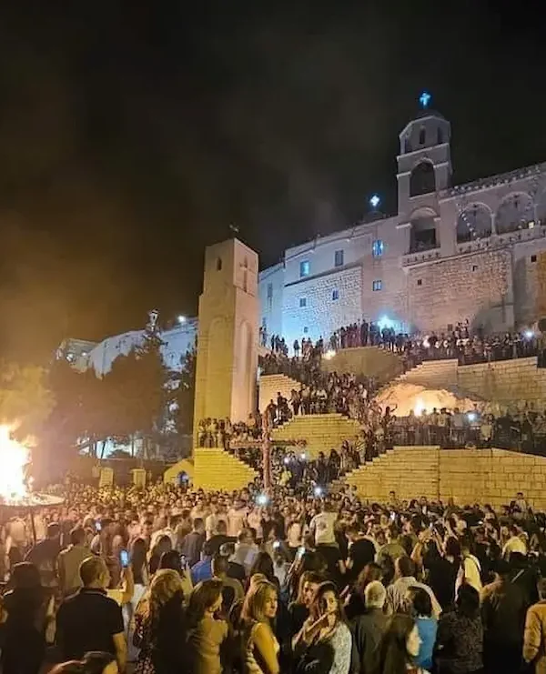 A large crowd of people gathered at night in front of the illuminated stone walls of the Our Lady of Saidnaya Monastery, watching a large ceremonial bonfire during a traditional religious festival.