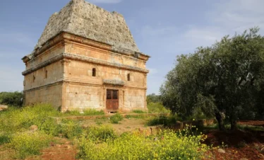 A well-preserved ancient stone tomb with a distinctive pyramidal roof standing in a field of yellow wildflowers and olive trees under a clear blue sky.