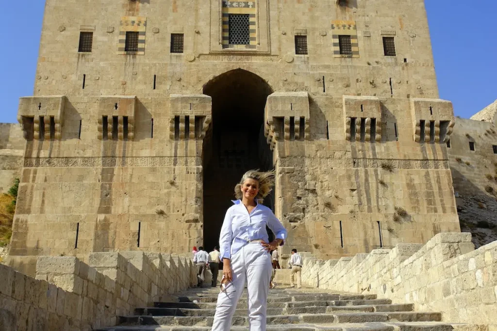 A traveler in white standing on the stone steps leading up to the massive, fortified gatehouse of the Aleppo Citadel, featuring intricate Islamic masonry and defensive arrow slits under a bright blue sky.