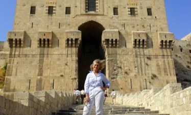A traveler in white standing on the stone steps leading up to the massive, fortified gatehouse of the Aleppo Citadel, featuring intricate Islamic masonry and defensive arrow slits under a bright blue sky.