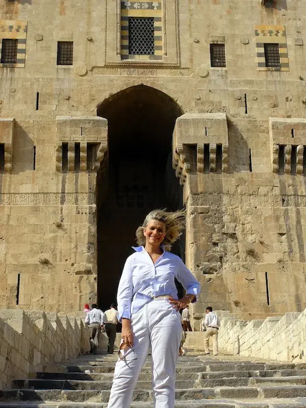 A traveler in white standing on the stone steps leading up to the massive, fortified gatehouse of the Aleppo Citadel, featuring intricate Islamic masonry and defensive arrow slits under a bright blue sky.