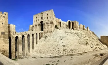 A wide panoramic view of the massive stone Citadel of Aleppo, showing the reinforced entrance bridge with high arches leading up to the main gatehouse atop a steep, fortified hill under a deep blue sky.
