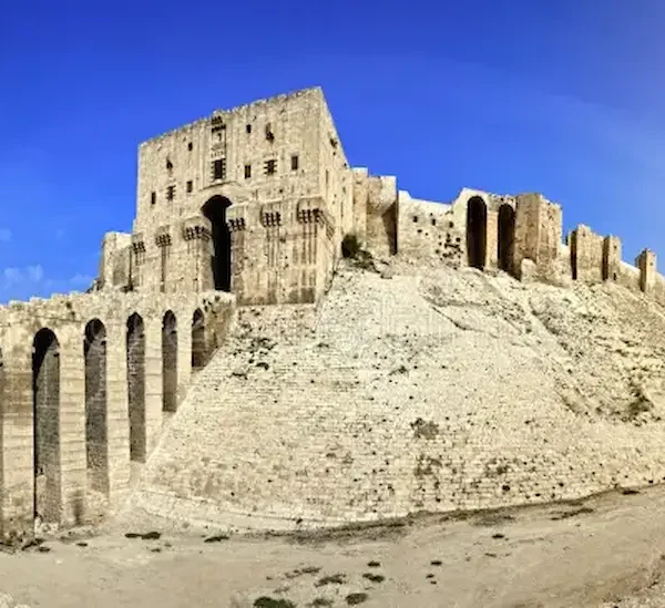 A wide panoramic view of the massive stone Citadel of Aleppo, showing the reinforced entrance bridge with high arches leading up to the main gatehouse atop a steep, fortified hill under a deep blue sky.
