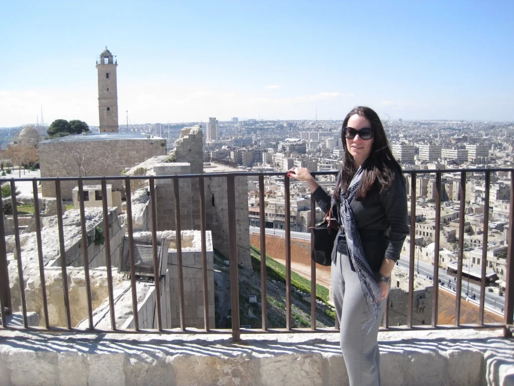 A woman with long dark hair and sunglasses stands at a metal railing on the Aleppo Citadel, with a sprawling cityscape, ancient stone walls, and a tall square minaret in the background under a clear sky.