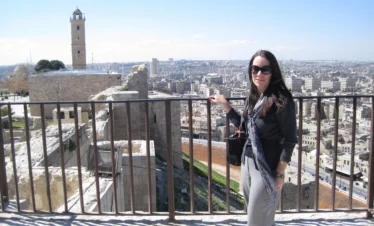 A woman with long dark hair and sunglasses stands at a metal railing on the Aleppo Citadel, with a sprawling cityscape, ancient stone walls, and a tall square minaret in the background under a clear sky.