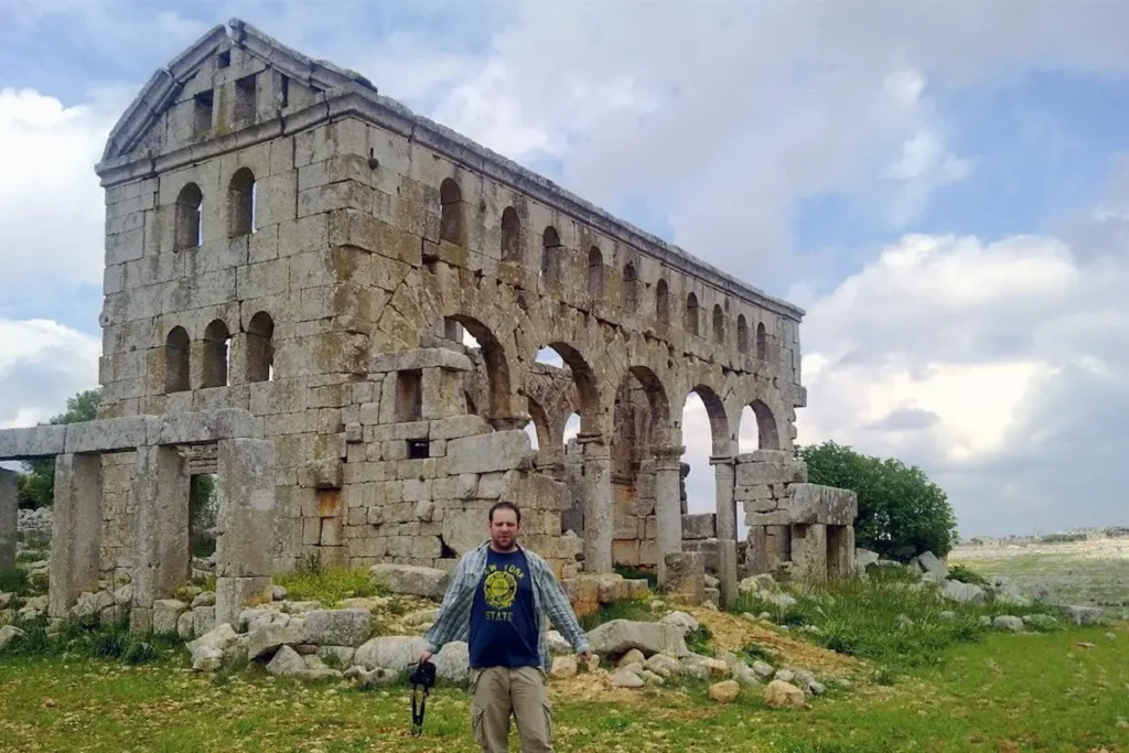 A traveler stands in a grassy field in front of the well-preserved ruins of a 5th-century stone church, featuring two levels of arched windows and a gabled roofline against a cloudy sky.