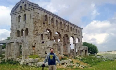 A traveler stands in a grassy field in front of the well-preserved ruins of a 5th-century stone church, featuring two levels of arched windows and a gabled roofline against a cloudy sky.