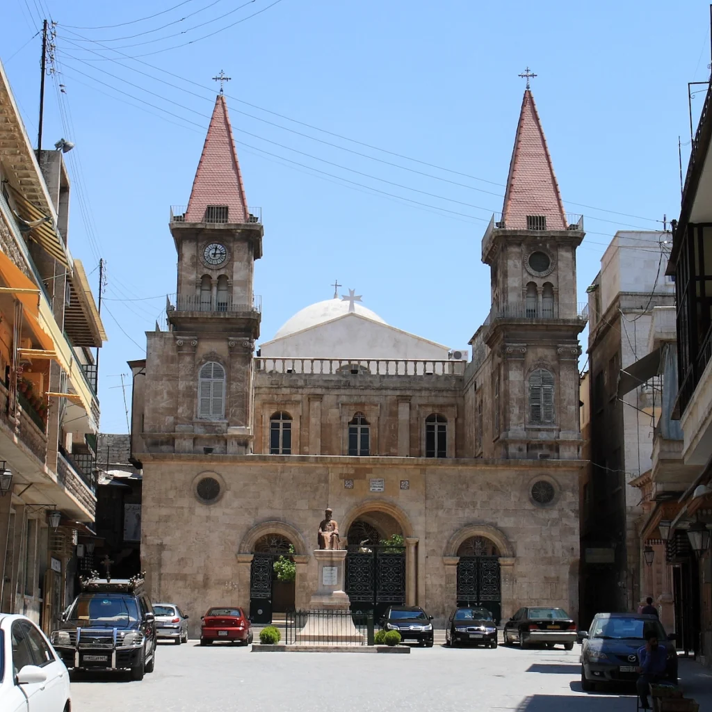 A grand stone cathedral with two symmetrical square bell towers topped with red pointed roofs, a central white dome, and three arched entrance gates located in a sunlit city square with parked cars.