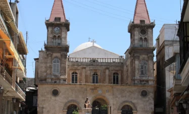 A grand stone cathedral with two symmetrical square bell towers topped with red pointed roofs, a central white dome, and three arched entrance gates located in a sunlit city square with parked cars.