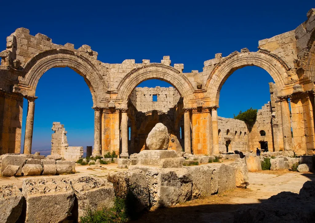 The sun-drenched ruins of the Church of Saint Simeon Stylites, featuring three massive limestone arches supported by columns, with the base of the saint's pillar sitting in the center under a deep blue sky.