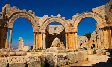 The sun-drenched ruins of the Church of Saint Simeon Stylites, featuring three massive limestone arches supported by columns, with the base of the saint's pillar sitting in the center under a deep blue sky.