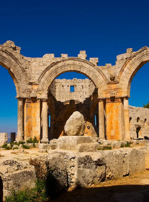 The sun-drenched ruins of the Church of Saint Simeon Stylites, featuring three massive limestone arches supported by columns, with the base of the saint's pillar sitting in the center under a deep blue sky.