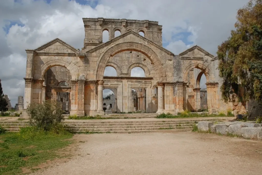 The large limestone facade of the ancient Church of Saint Simeon Stylites in Syria, featuring three arched entrances, classical columns, and a central pediment under a cloudy blue sky.
