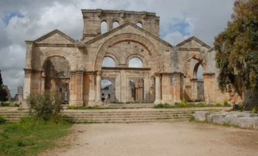 The large limestone facade of the ancient Church of Saint Simeon Stylites in Syria, featuring three arched entrances, classical columns, and a central pediment under a cloudy blue sky.